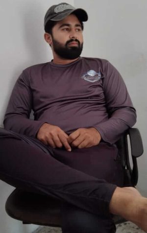 Bearded Man in a Cap Sitting Cross-legged in an Office Chair Against a Gray Wall, Looking to the Side and Relaxed.