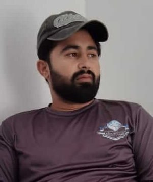 Bearded Man in a Cap Sitting Cross-legged in an Office Chair Against a Gray Wall, Looking to the Side and Relaxed.